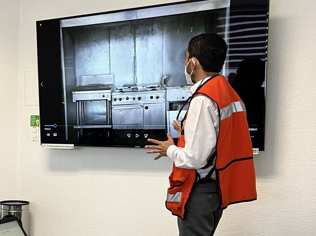 A specialist in a high-visibility vest presents a video of an industrial kitchen on a wall-mounted screen.