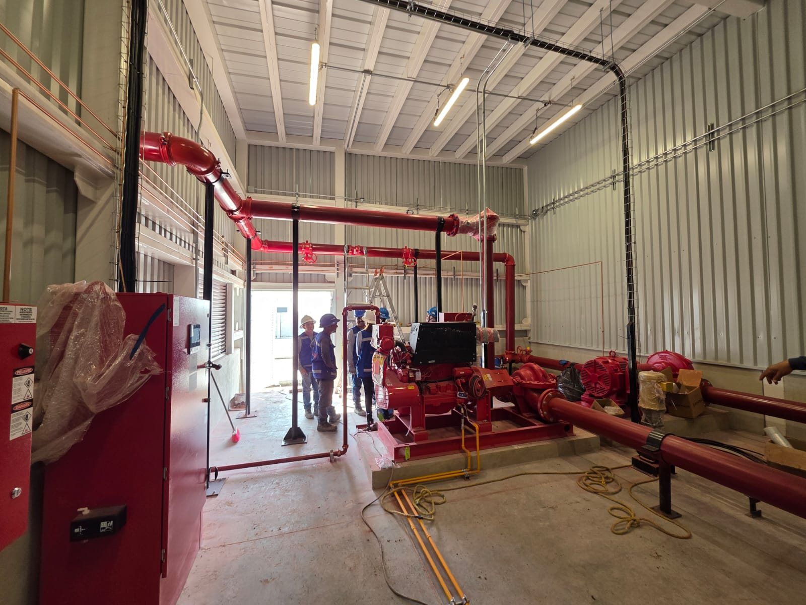Fire pump room in a sheet-metal industrial building, with the red piping and the pumping equipment installed.