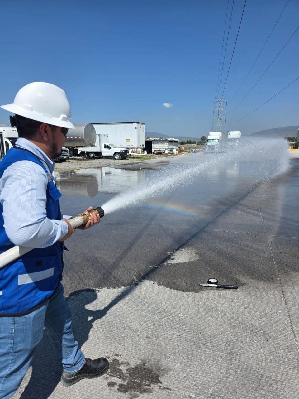 A technician in a white hard hat directs a hose stream during a test in a plant yard; on the ground, a pitot tube with a pressure gauge.