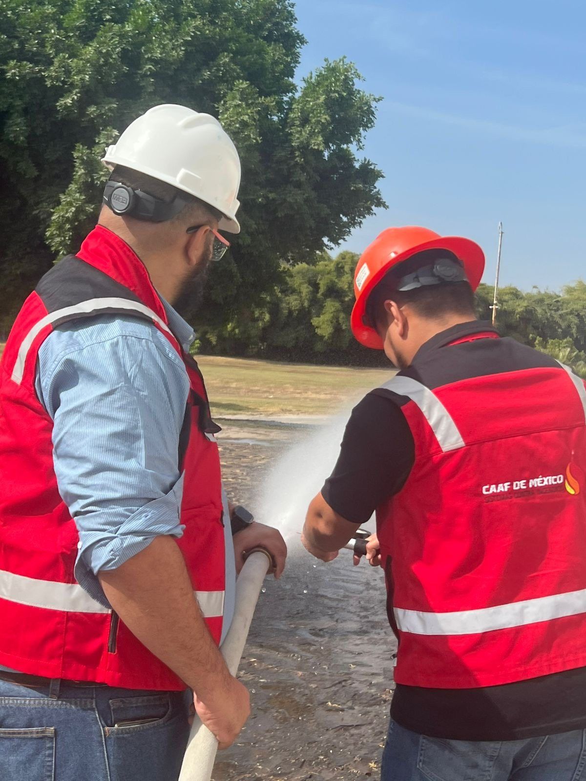 Two CAAF de México technicians in red vests and hard hats hold a hose discharging pressurized water.