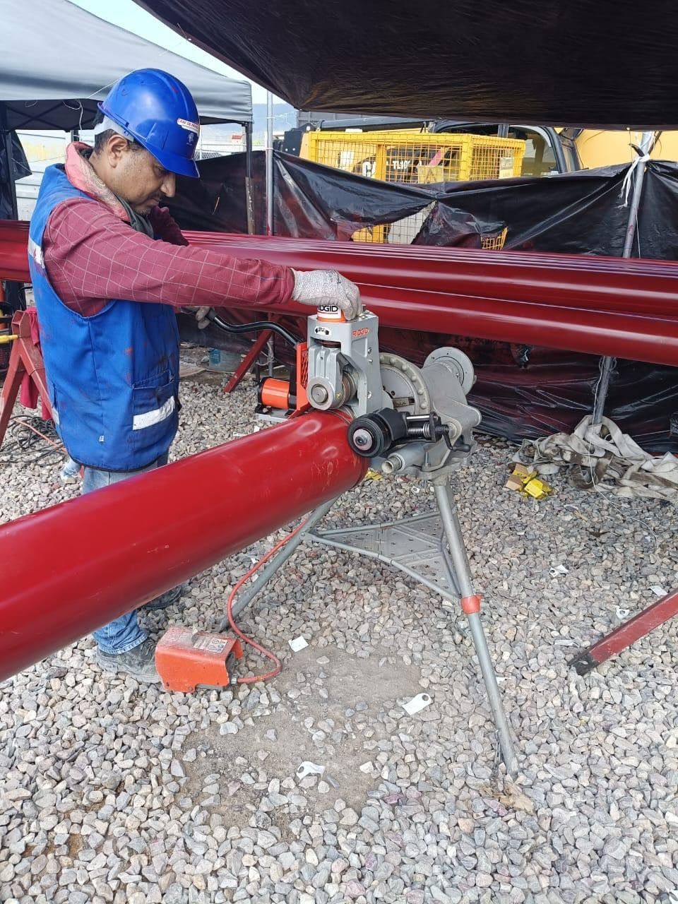 A technician grooves a length of red steel pipe with a pipe groover mounted on its tripod, in the yard of a job site.